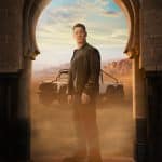 A man in a black leather jacket stands in an arched doorway with desert mountains and an off-road vehicle behind him, dust swirling around his feet.