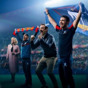 Four people cheer at a sports stadium, two men in front celebrating with arms raised and flags, while a man and woman in coats stand behind them amid bright stadium lights—a scene reminiscent of the energetic spirit seen in an Apple TV comedy like Ted Lasso.