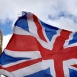 The Union Jack flag waves in the foreground with Big Ben and part of the Houses of Parliament visible against a cloudy sky in London, England—a fitting backdrop as debates over UK App Store rules and price regulation shape the nation’s digital landscape.