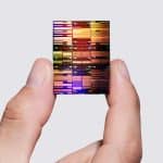 A hand holds a colorful, reflective 0.7nm chip between the thumb and index finger against a plain, light gray background.