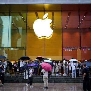 A crowd with umbrellas stands outside a large Apple Store with a glowing Apple logo on the glass facade. It appears to be raining, and people are gathered near the store entrance—a scene that highlights the brand’s immense popularity in the China market.