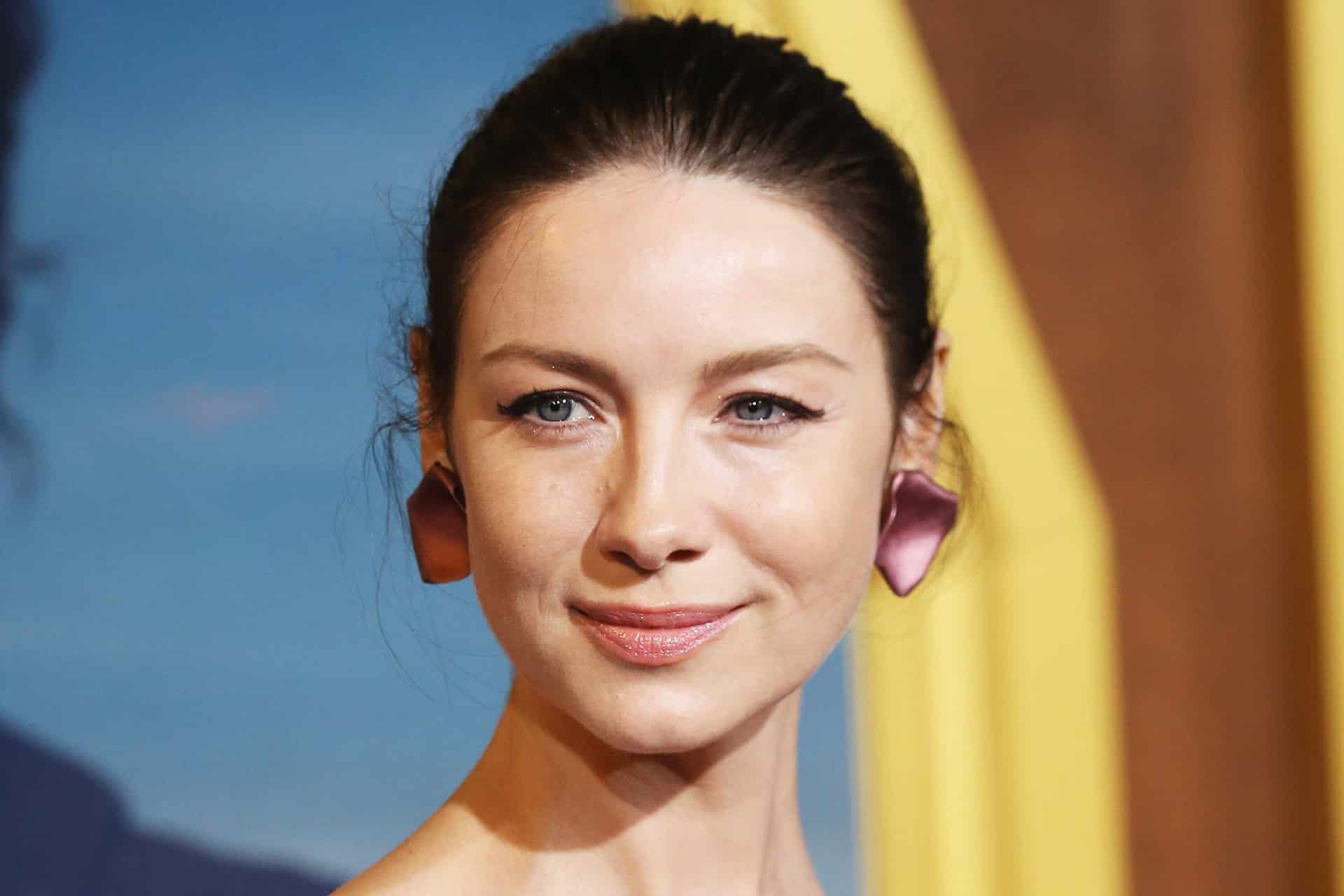 A woman with fair skin, blue eyes, and dark brown hair pulled back, wearing mauve, petal-shaped earrings, smiles softly in front of a blurred blue and yellow background—her serene presence reminiscent of the thoughtful storytelling found in Alfonso Cuarón’s acclaimed works and the immersive visuals seen throughout Apple TV’s Ascension series.