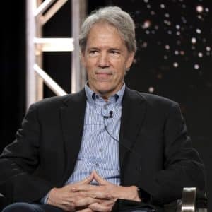 An older man with gray hair sits in a chair, wearing a dark suit and striped shirt. He has his hands folded and a microphone clipped to his shirt. The background features lights and a metal scaffolding, evoking the atmosphere of a Warner Bros set or an Apple TV production.