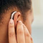 Close-up of a person’s hand adjusting a behind-the-ear hearing aid placed on their ear, with a blurred background. The device is one of the latest Made for iPhone (MFi hearing devices), ensuring seamless connectivity with Apple hearing aids technology.