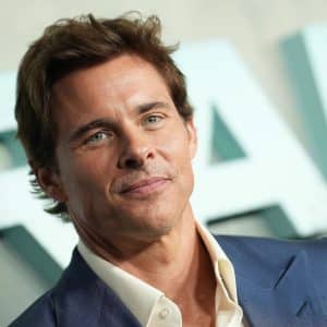 A man with short brown hair, wearing a blue suit jacket and white shirt, smiles slightly while standing in front of a blurred background with large, light-colored letters. The subtle confidence in his expression might remind viewers of James Marsden’s roles on popular platforms like Apple TV.