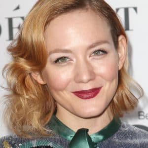 Louisa Krause - A woman with wavy blonde hair and red lipstick smiles at the camera, wearing a green and blue outfit, standing in front of a white background with partially visible black text—reminiscent of Dakota Fanning’s recent appearance at an Apple thriller premiere.