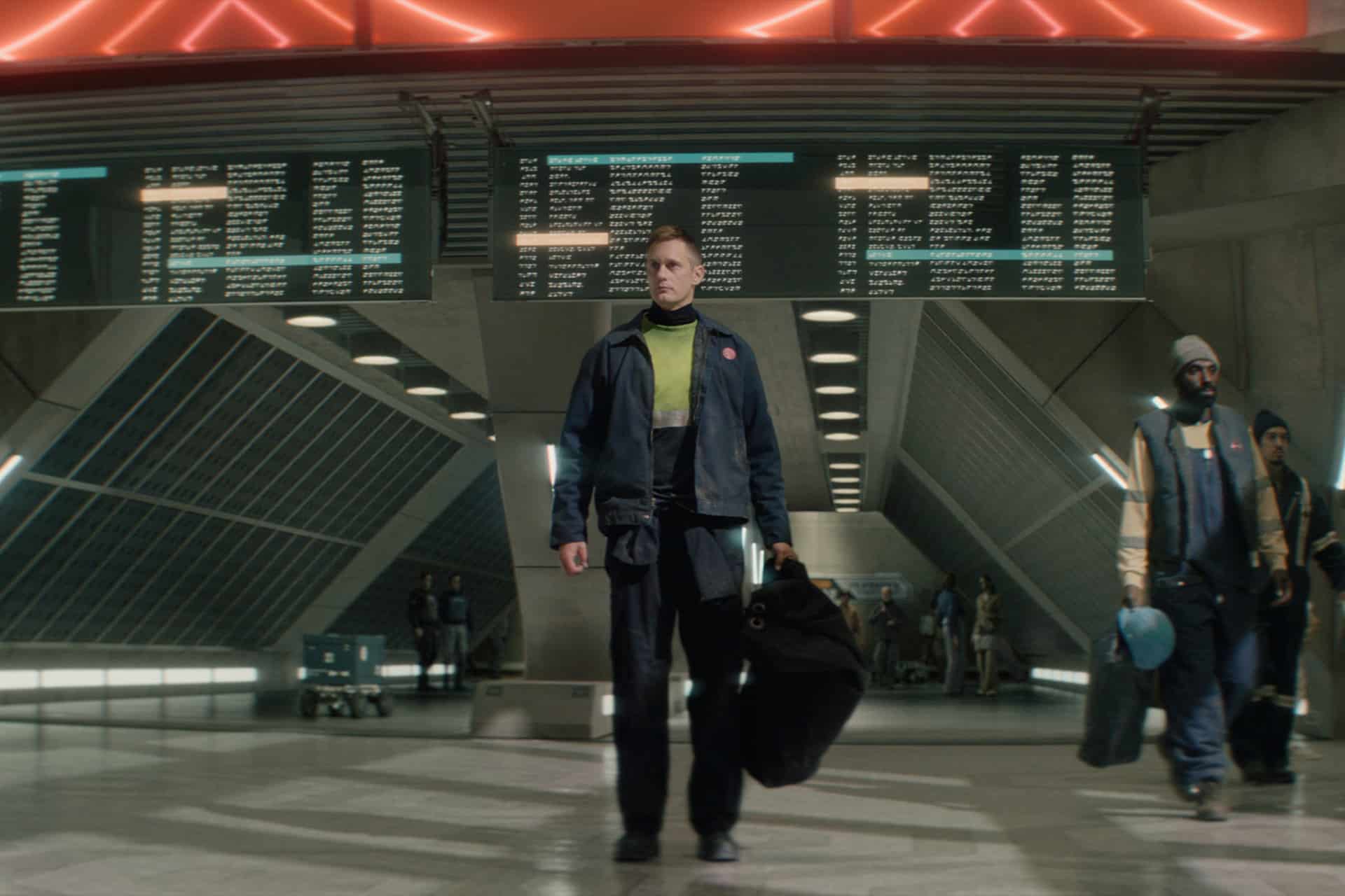 A person carrying bags stands in a modern, futuristic station beneath large digital departure boards, with other travelers visible in the background—a scene reminiscent of the sleek settings from Murderbot Season 2 on Apple TV.