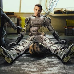 A man in a damaged, futuristic robotic suit—reminiscent of Murderbot from Apple TV’s Murderbot Season 2—sits on the floor with wires exposed and connected to a robotic arm. The background shows a sci-fi interior with yellow containers and round windows.