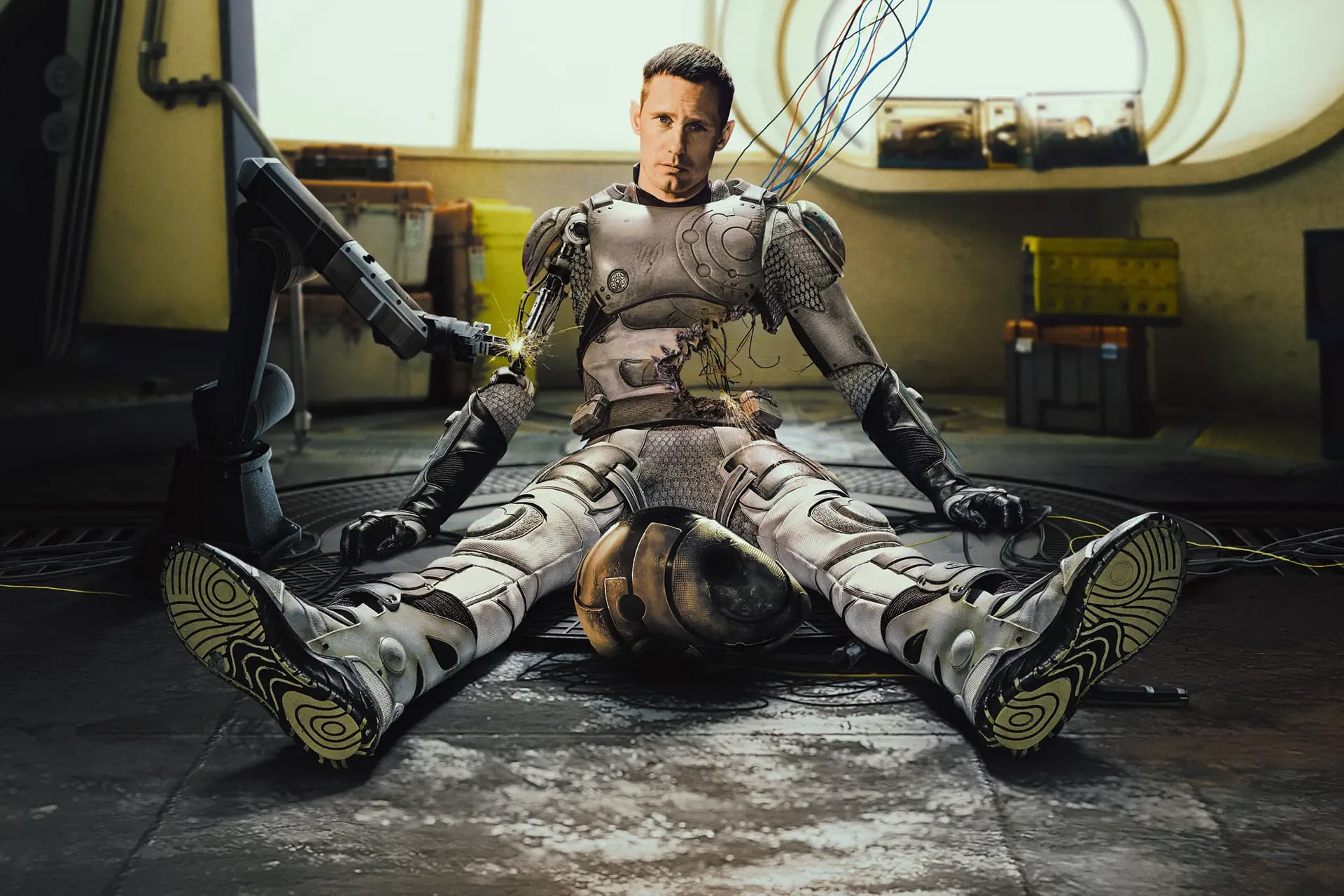 A man in a damaged, futuristic robotic suit—reminiscent of Murderbot from Apple TV’s Murderbot Season 2—sits on the floor with wires exposed and connected to a robotic arm. The background shows a sci-fi interior with yellow containers and round windows.