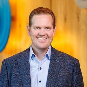 A man in a blue checked blazer and light blue shirt smiles, standing in front of a wooden wall with large colorful circular art pieces—evoking the professional confidence often seen among Apple leadership and experts in government affairs.