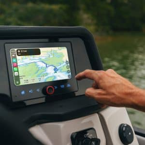 A person’s hand touches a marine navigation GPS screen on a boat dashboard, showing a route over a waterway. Green trees and water are visible outside, highlighting the perfect setting for cruising on pontoon boats.