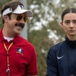 A man in a red polo shirt and visor smiles, standing next to a serious-looking woman in a navy jacket. Both have logos on their clothing—one of which hints at Apple TV, perhaps nodding to a streaming record set by Jason Sudeikis’s beloved sports series. They are outdoors with blurred trees in the background.