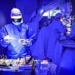 Surgeons in blue gowns perform an operation in a room lit with blue light. One surgeon stands in the foreground handling instruments, while others operate on a patient in the background. Advanced technology is present, as this state-of-the-art operating room integrates Apple Vision Pro, enhancing precision throughout the procedure. Surgical tools are arranged on a tray.